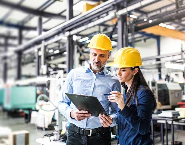 A man and woman wearing hardhats looking at a clipboard in a manufacturing facility